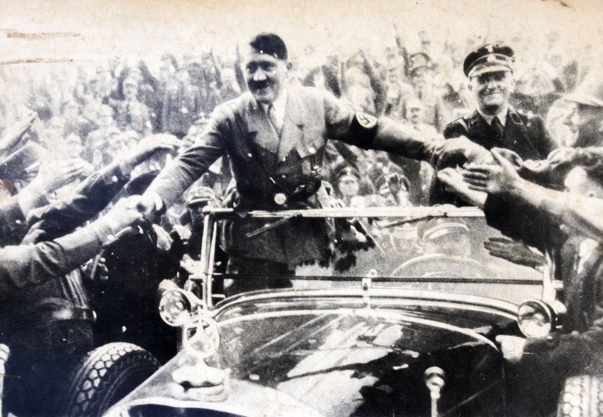 Adolf Hitler stands in a convertible and shakes hands with his followers