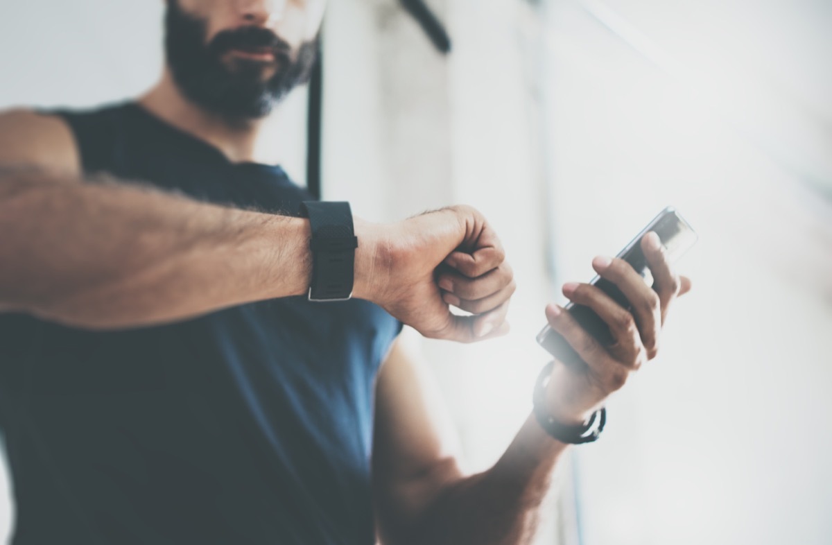Close-up Shot Bearded Sportive Man After Workout Session Checks Fitness Results Smartphone.Adult Guy Wearing Sport Tracker Wristband Arm.Training hard inside gym.Horizontal bar background.Blurred - Image