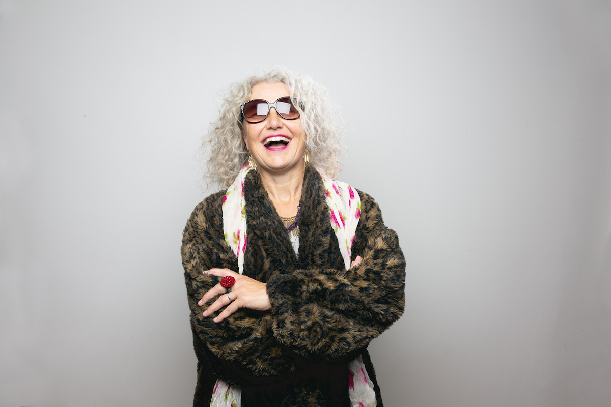 Cool mature woman, wearing sunglasses and a coat made from artificial fur, wearing scarf with floral pattern and a lot of jewelry, standing in front of grey background