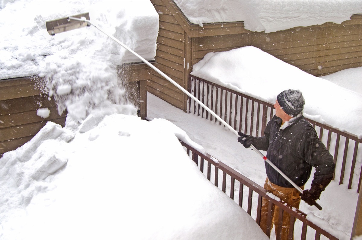 white man in winter clothing raking snow off roof