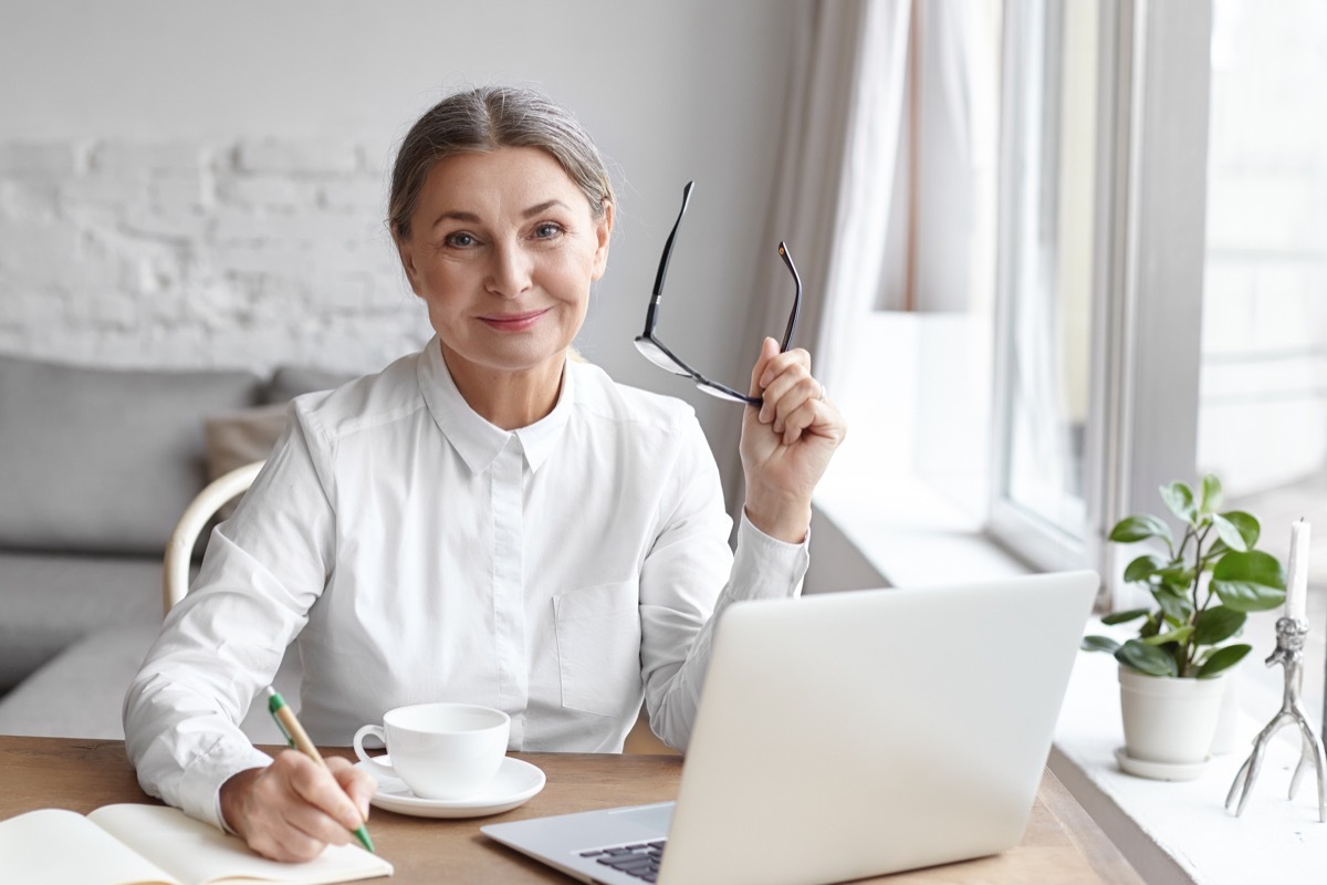 A mature woman wearing a white blouse sits at her desk next to a window writing in a pad and using her laptop