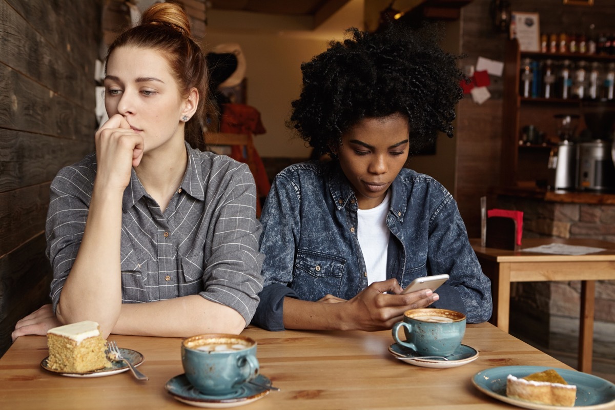 two women sitting at cafe; one uses her phone while the other looks away annoyed