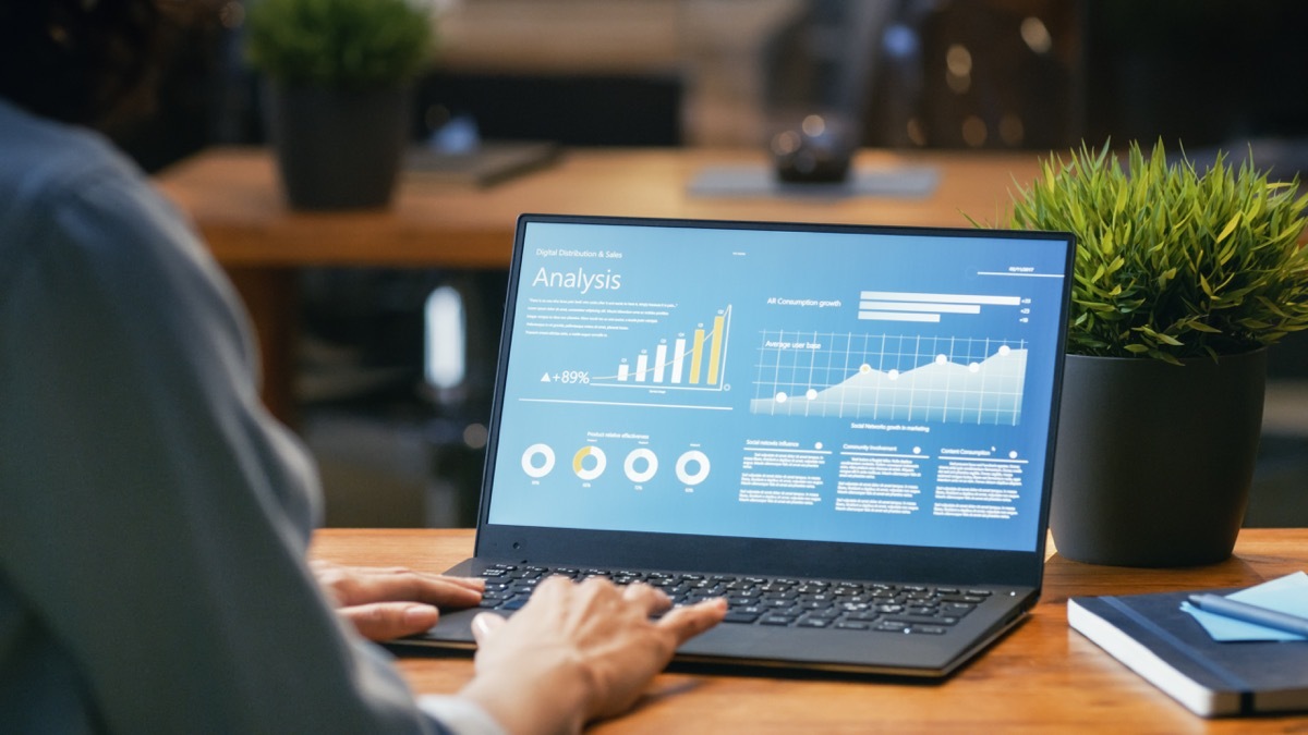 Female Analyst at Her Desk Works on a Laptop Showing Statistics, Graphs and Charts. She Works on the Wooden Table in Creative Office. Over the Shoulder Footage.