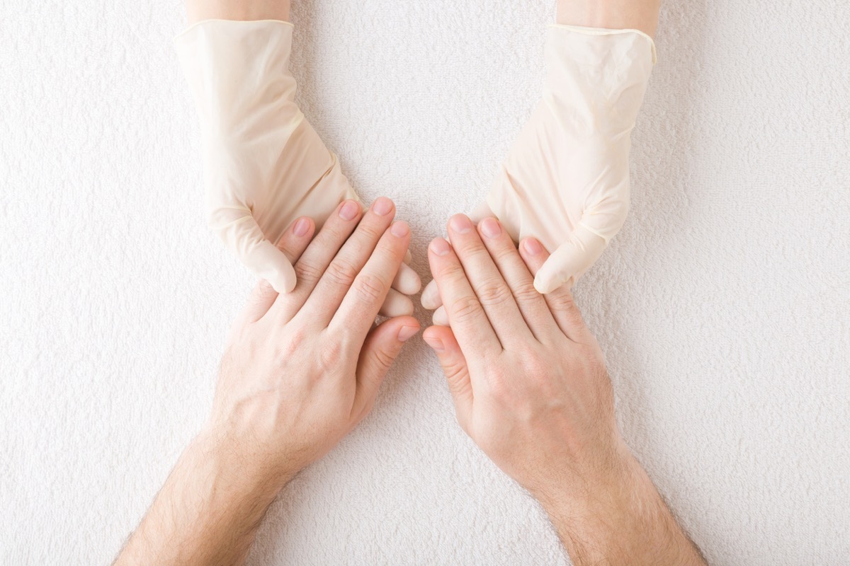 Manicurist hands in rubber protective gloves holding young adult man fingers on white towel background. Manicure, pedicure beauty salon concept. Closeup. Care about fingernails. Top down view.