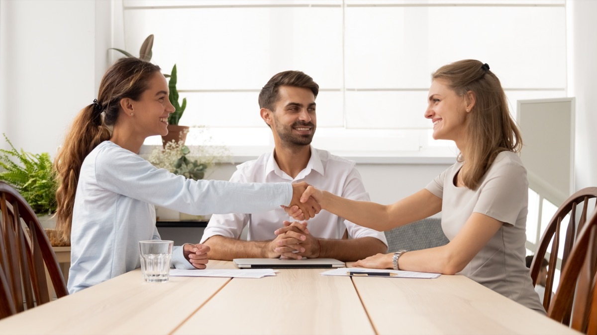 Two females smiling and shaking hands with a male mediator sitting at the table between them