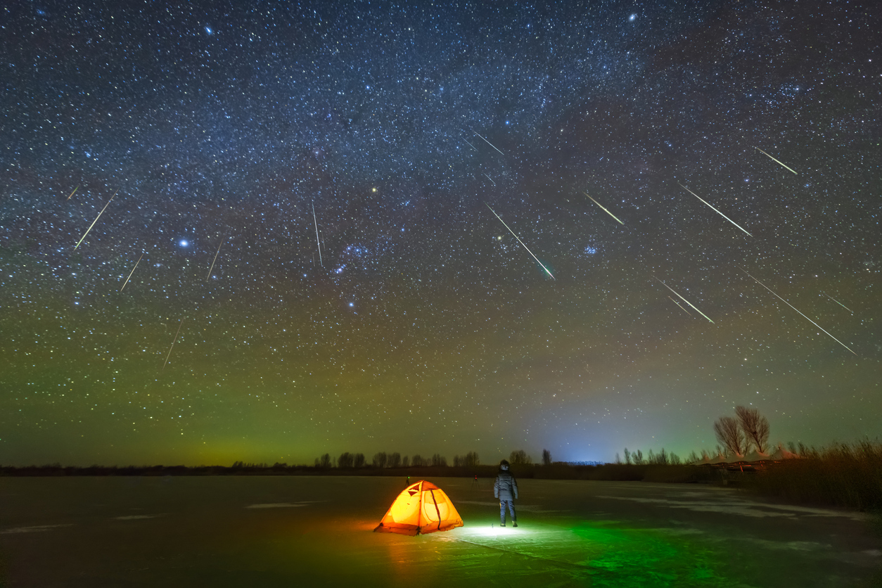 A person standing outside their tent looking up at shooting starts during a meteor shower