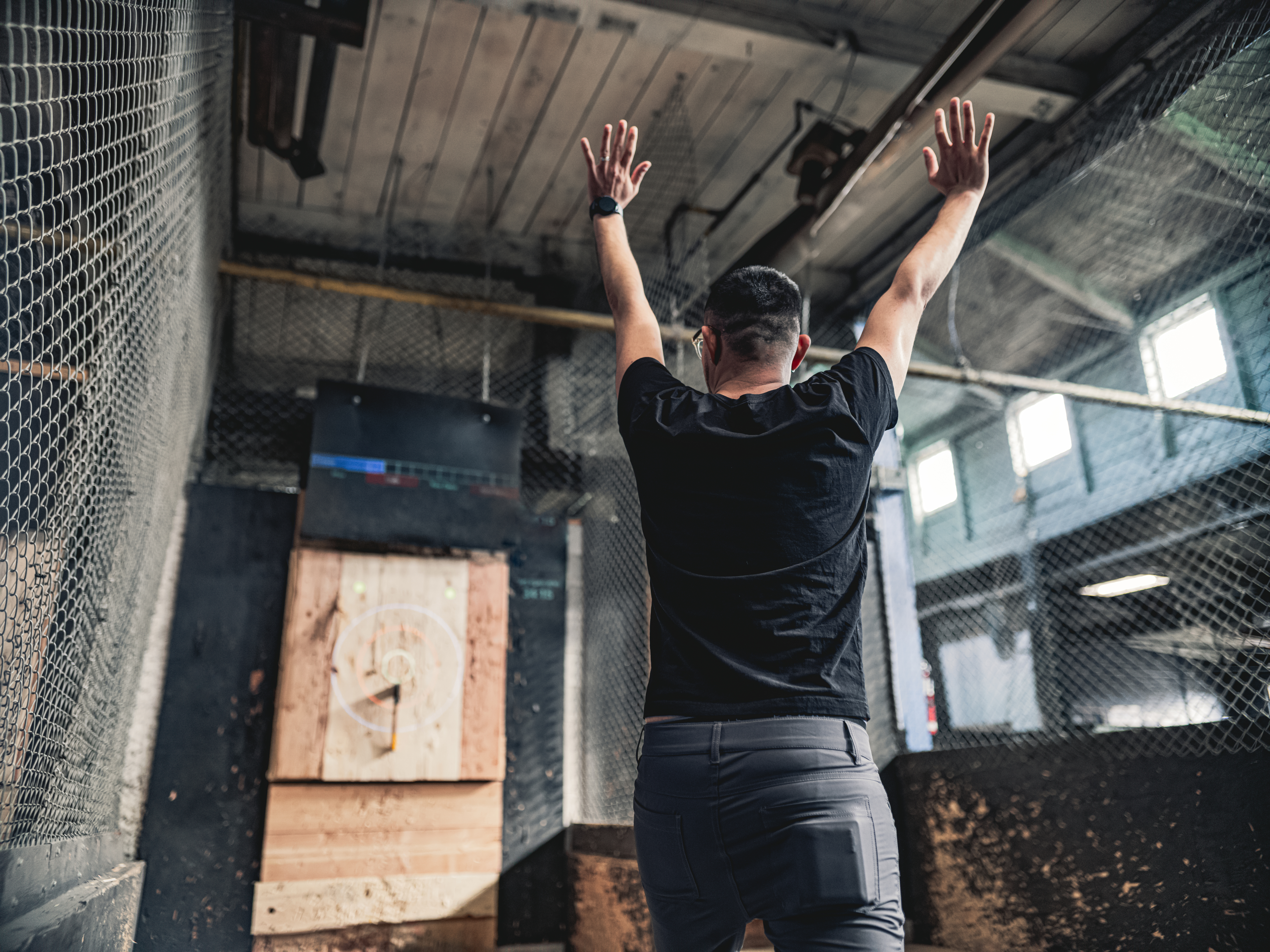 Young Middle Easter gay man throwing axe at the game range. He is dressed in casual clothing, wearing eyeglasses. Interior of warehouse like space during the day.