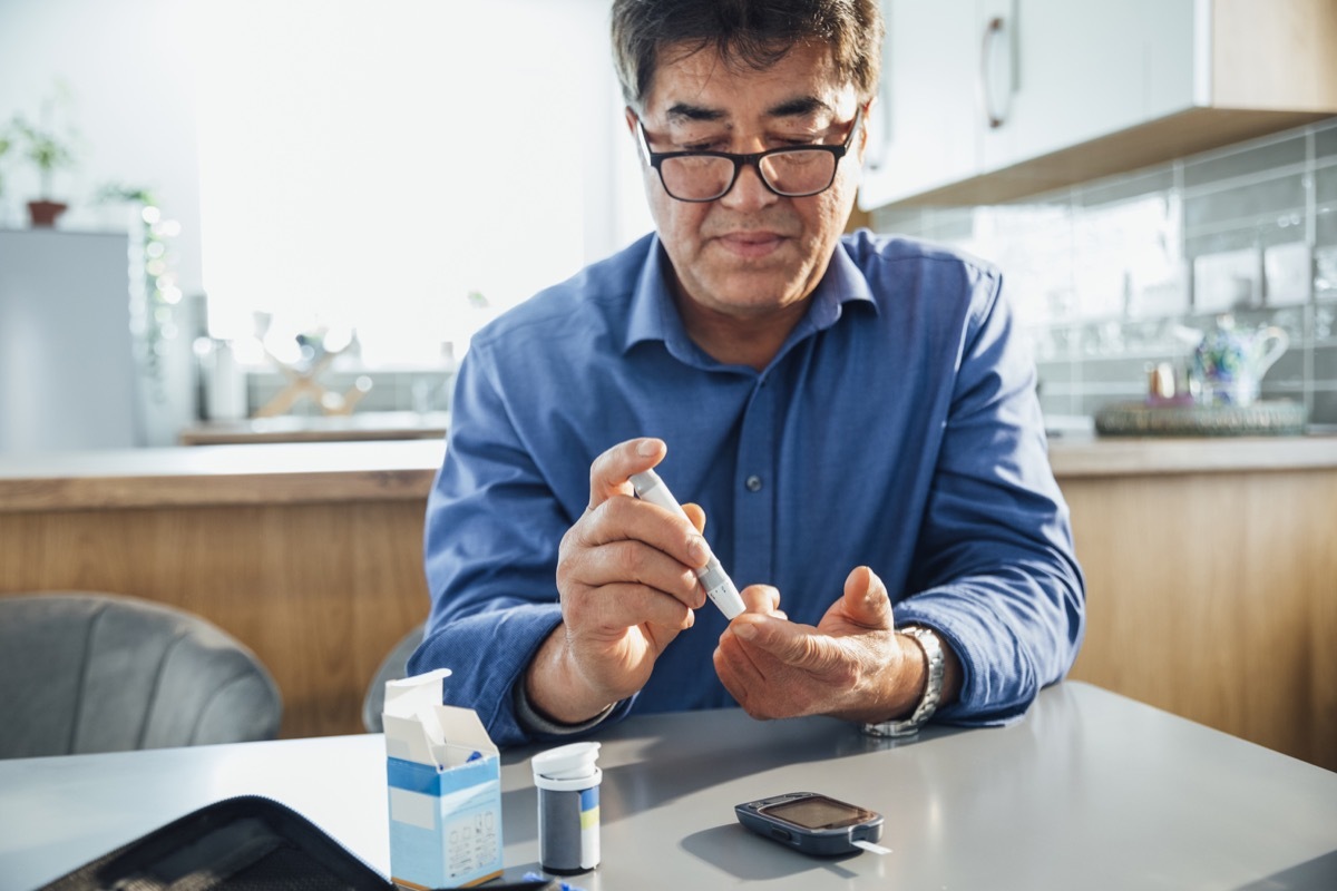 man sitting at a dining table in his kitchen, he is pricking his finger using a glaucometer to test his blood sugar levels, he is managing his diabetes.