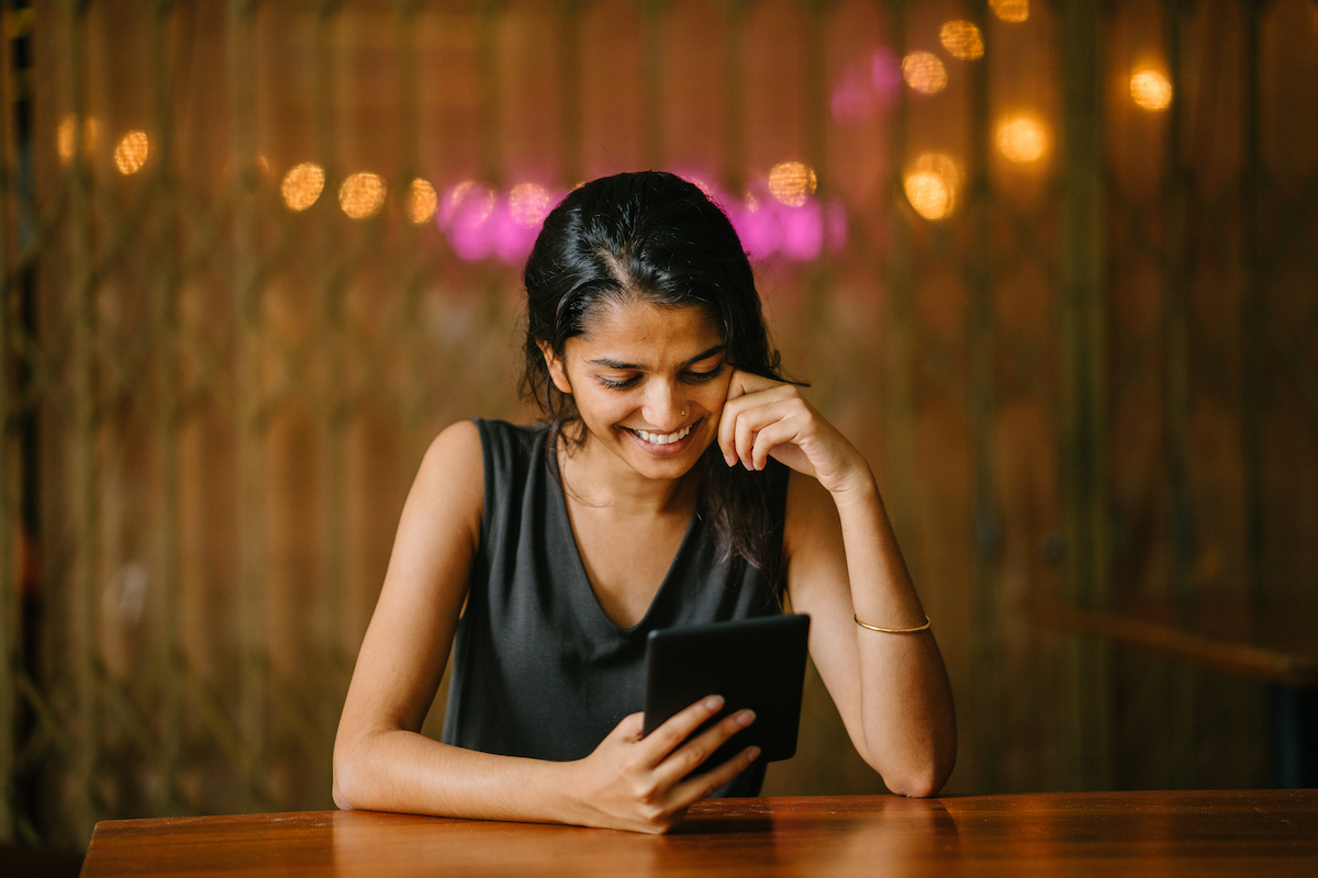 woman in a black blouse is casually relaxing and reading a tablet e reader (e.g. a Kindle) at a wood table in a warm, cozy setting.