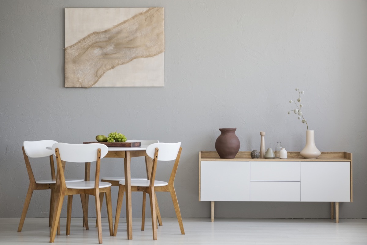 Real photo of wooden table and four chairs standing in light grey dining room interior with modern art poster and cupboard with decor
