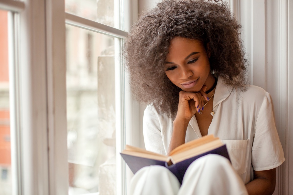 A young woman in white pajamas reading a book on a window seat