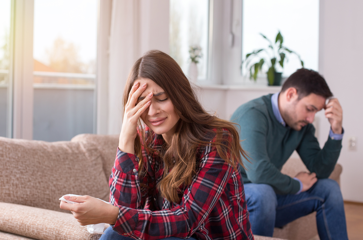 A young couple facing away from each other on the couch; the man has his head in his hands and the woman is crying.