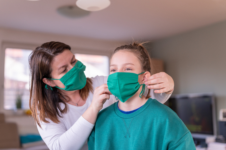 A mother wearing a green homemade protective face mask puts the same style of mask on her daughter