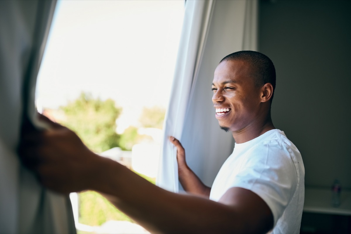 Shot a handsome young man opening his bedroom curtains in the morning