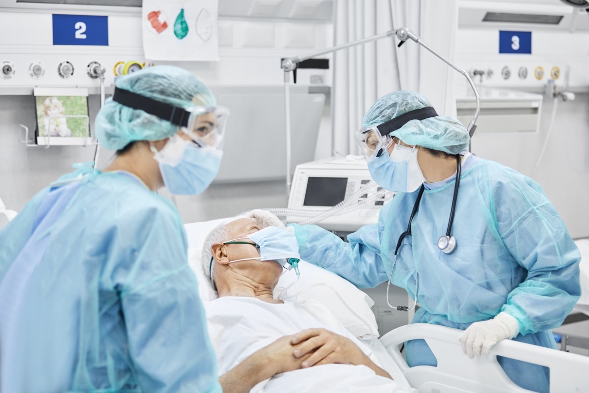 Female doctor talking to a patient who is lying on hospital bed amidst essential workers. 