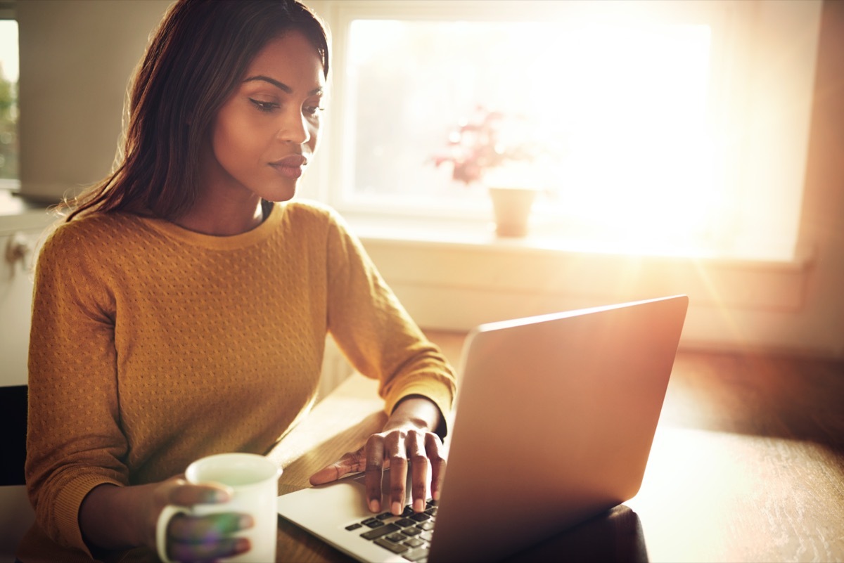 female sitting at table holding coffee cup and typing on laptop with light flare coming through window